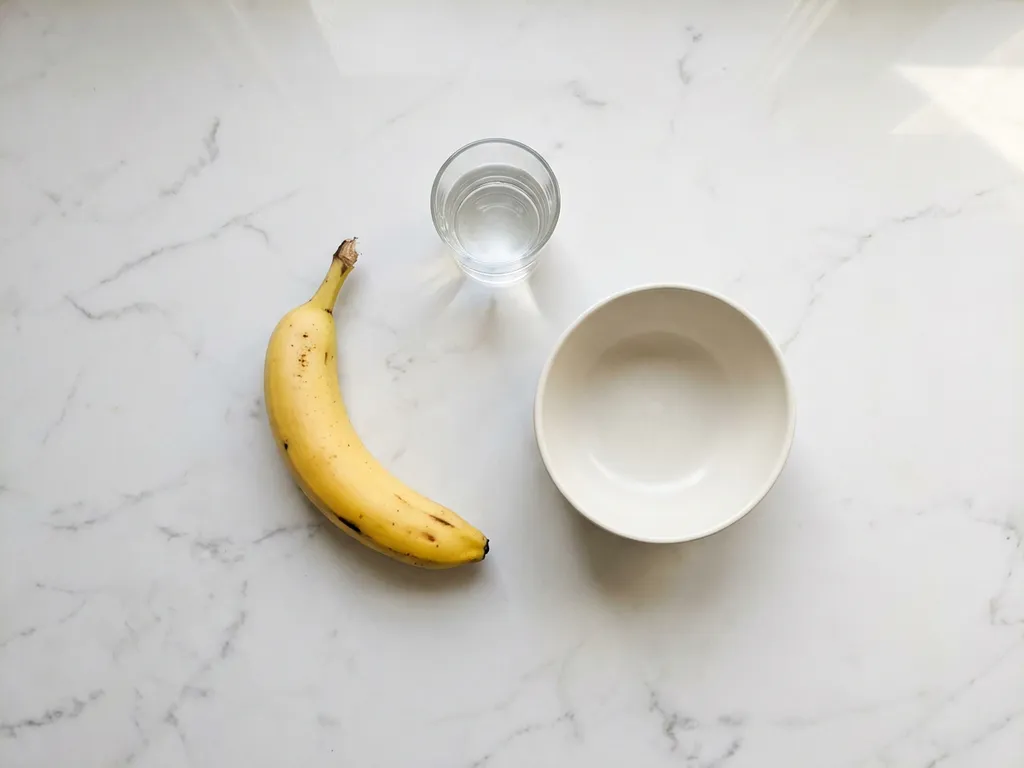 Ripe yellow banana on white marble surface with glass of water and ceramic bowl in minimalist flat lay composition