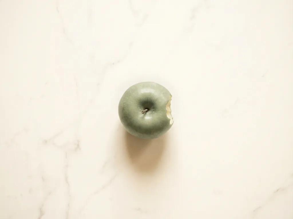 Green apple with bite taken out sitting on white marble surface with soft natural lighting and shadows
