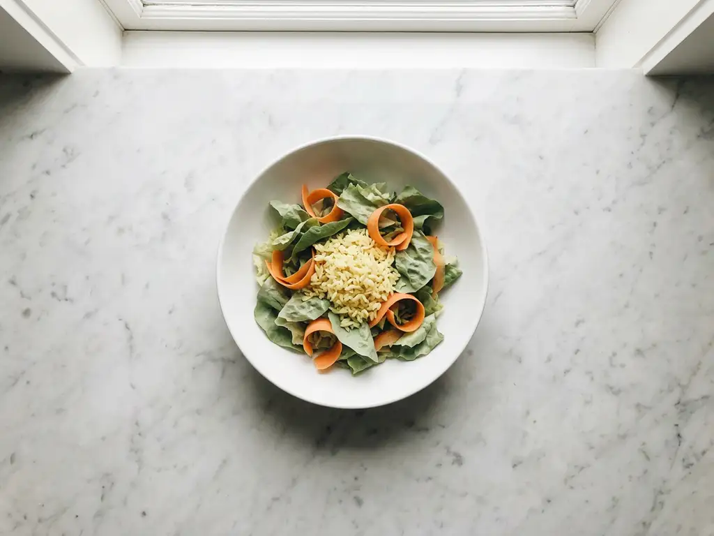 White ceramic bowl with lettuce leaves, carrot ribbons, and yellow rice on marble countertop, overhead view with soft lighting
