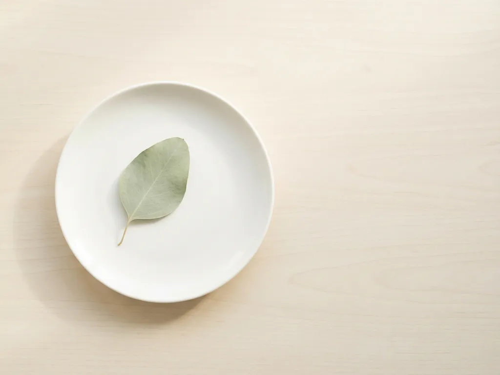 Single pale green leaf on white ceramic plate with soft shadows on light wooden table surface