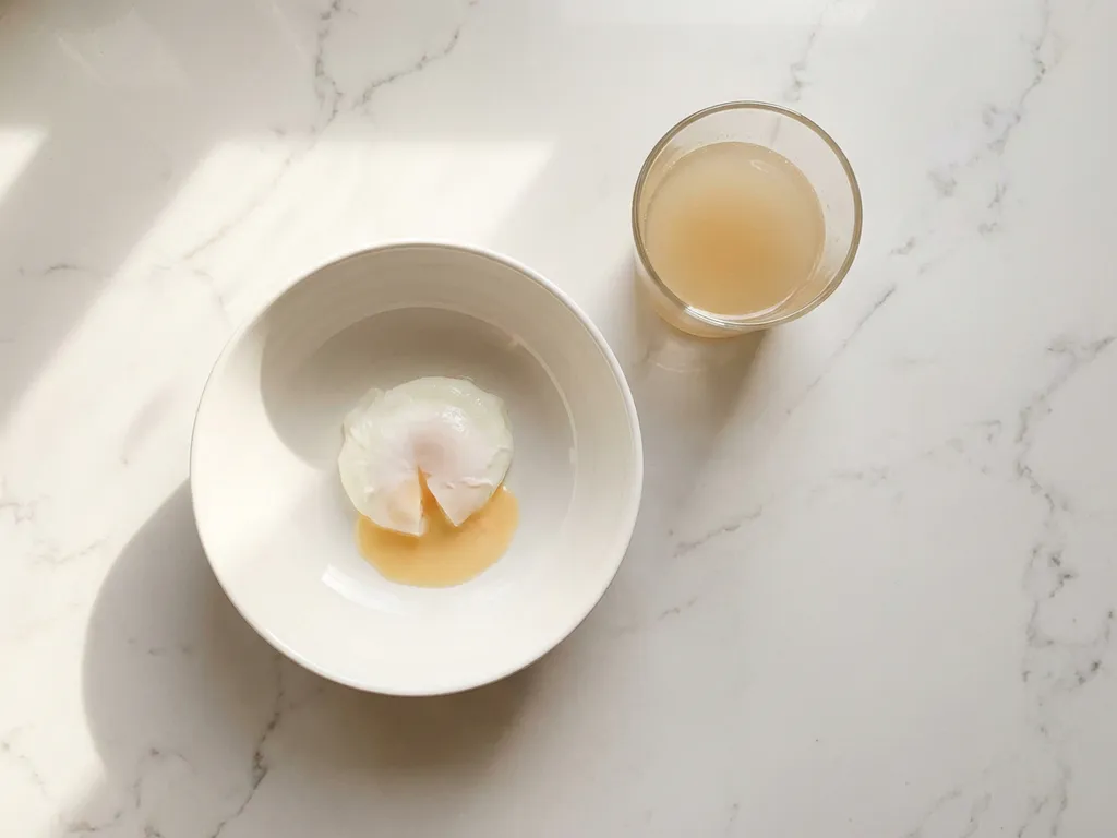 White ceramic bowl with poached egg and runny yolk on marble surface next to glass of bone broth, overhead view